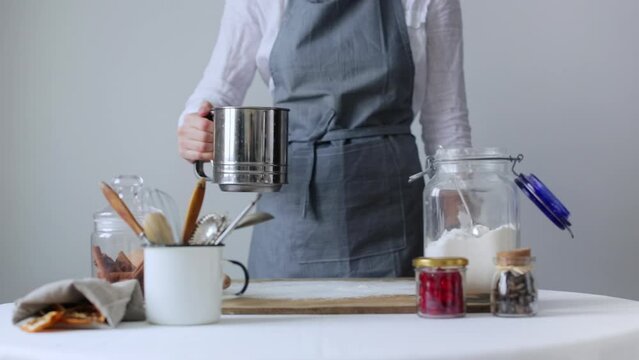 Woman baker in grey apron sifting flour with flour sifter on a wooden board for making dough, preparing dough for baking cake, bread, dessert, cookies. Get rid of lumps with the Baking Sifter Tools