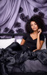 Naklejka premium Beautiful brunet woman with curly hair posing on her bed, purple background. 