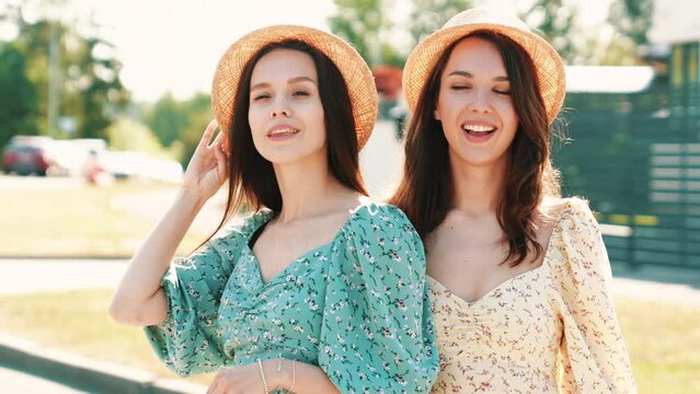 Two young beautiful smiling hipster woman in trendy summer sundress. Carefree women posing in street in hats. Positive models having fun and hugging at sunset outdoors. Cheerful and happy