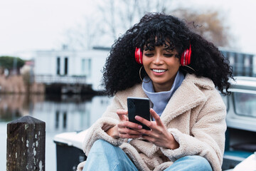 Smiling teenager with Afro hair listens favorite music track holds mobile phone downloads song to her playlist dressed in winter clothes. People hobby technology