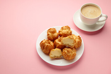 Homemade profiteroles with cream on plate with cup of coffee on pink background. Fresh baked golden profiterole.