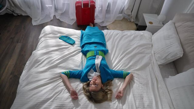 Top view gorgeous slim woman falling back on bed in slow motion in hotel room. Portrait of beautiful Caucasian stewardess enjoying leisure in bedroom indoors