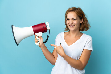 Young Georgian woman isolated on blue background holding a megaphone and pointing side