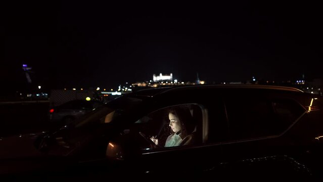 Beautiful woman browsing a smartphone, sitting in a car overlooking the night neon light city 