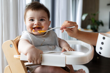 Cute baby sitting in highchair eating food pumpkin from spoon with mom