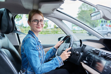 Teenage girl driver in glasses sitting behind wheel of car, looking at camera