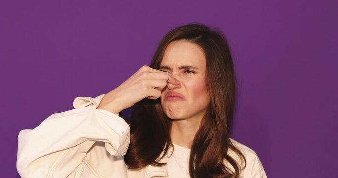 Disgusting odor. Discontent brunette curly woman frowning face, smells something awful, closing nose because of a bad smell, feeling aversion, purple studio background.