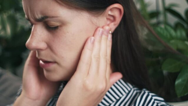 Close up of upset young female holding painful ears, suddenly feeling strong ache. Unhealthy sad Caucasian woman suffering from painful otitis sitting on couch at home. Health problems concept