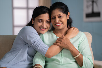 Happy indian mother hugging her daughter by looking camera while sitting on sofa at home - concept of Tender or Affection, Family Togetherness and Caring Connection.