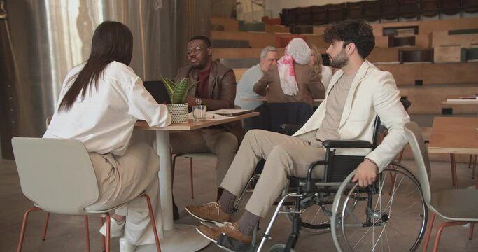 Businessman in wheelchair coming to the table for discussion with his inclusive colleagues in the office cafe bar area, trucking movement