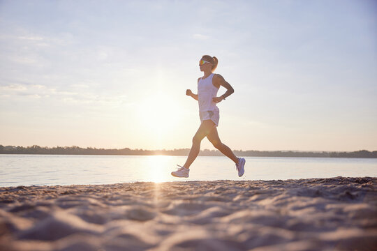 Side view of fitness woman running on beach by sea. Sportswoman training, warming up on seaside evening promenade at sunset.