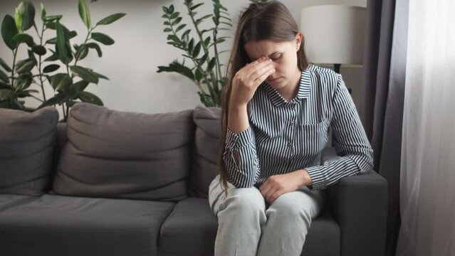 Unhappy depressed young caucasian girl hiding face in hand, sitting on couch alone in living room at home. Desperate millennial woman suffering from personal life problems or stress, headache concept