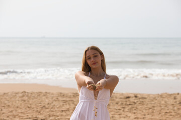 Fototapeta na wymiar Pretty young blonde woman in white dress spills sand from between her hands as she holds her hands out in front of her. The woman is happy and having fun. In the background the blue sea.