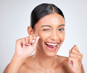 Woman, portrait and wink to floss teeth in studio for healthy dental care, gum gingivitis or plaque on white background. Face, happy indian model and oral thread for cleaning mouth, tooth and hygiene