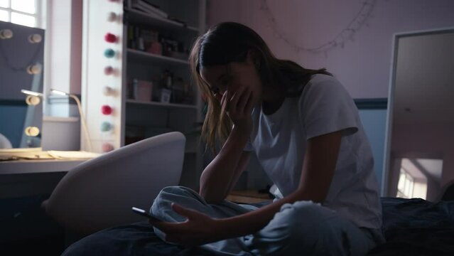 Depressed teenage girl sitting on bed at home looking at bullying messages on mobile phone - shot in slow motion
