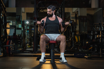 Fototapeta na wymiar handsome muscular caucasian man concentrated lifting dumbbell weight sitting on bench at modern gym, active fitness people workout functional training for body and mental health in dark sport club