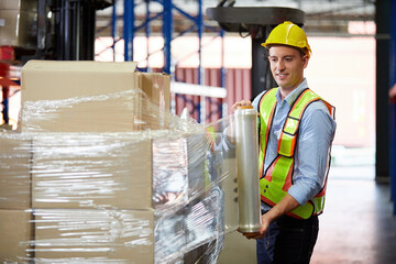 factory worker or warehouser wrapping stretch film parcel on pallet covered cardboard boxed in...