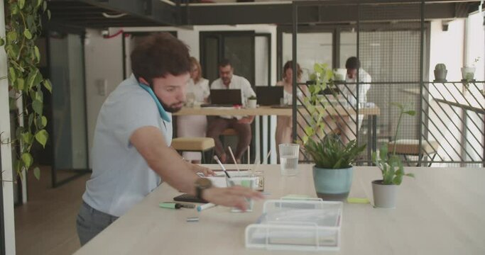 Caucasian male manager working at his desk, angry speaking on the phone while his colleagues are working together in the background in the office during summer time