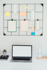 Modern workplace - white desk with laptop mockup empty screen, grid mood board with pinned notes and check lists for plans, office supplies at work space in home office room interior. Vertical card.