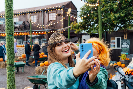 Two laughing female friends in fun glasses and witch hat having fun and making selfie on phone on decorated pumpkin farm. Selecting Thanksgiving and Halloween holidays decor. Autumn fall festive mood.