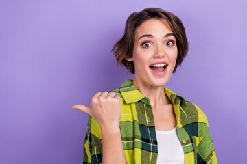Portrait of astonished young surprised girl directing finger empty space wear checkered shirt best offer isolated purple color background