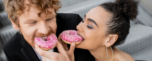 elegant interracial couple feeding each other with sweet donuts, wedding on street, fun, banner