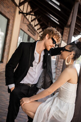 stylish groom in sunglasses, with hand in pocket near elegant african american bride on urban street
