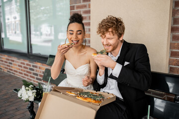 smiling african american bride and redhead groom eating pizza on bench, wedding celebration in city