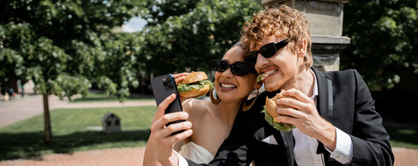 interracial newlyweds in sunglasses eating burgers and taking selfie on smartphone on street, banner