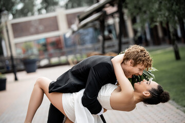 Fototapeta premium young excited man embracing elegant african american bride on street of european city, side view