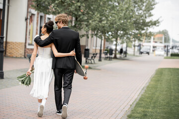 back view of multiethnic elegant newlyweds with bouquet and longboard embracing and walking in city - Powered by Adobe