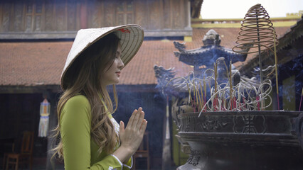 Portrait of Asian Vietnamese woman with Vietnam dress and straw hat praying by incense in a temple.