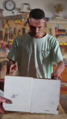 Young carpenter apprentice working at carpentry workshop with drilling tool repairing furniture wood with the supervision of mentor in vertical video