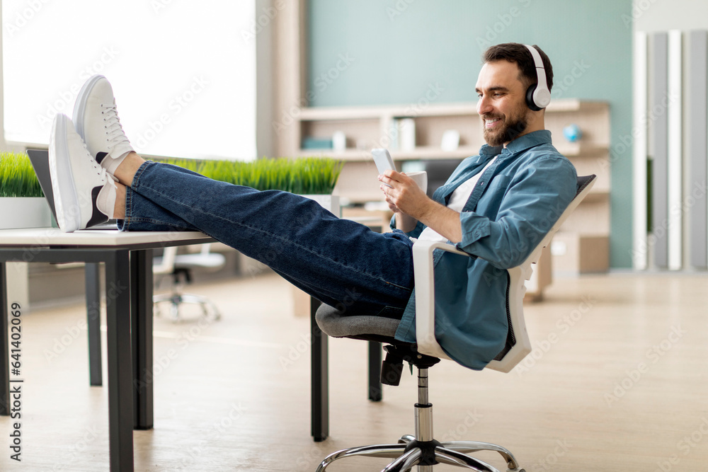 Poster Smiling man relaxing in office, resting feet on table and using smartphone