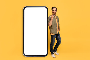 Cheerful middle aged man leaning and pointing at big smartphone with white blank screen, smiling at camera, mockup