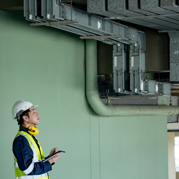 Building inspector man checking telephone cable duct wireway. Asian male maintenance worker with green vest and safety helmet pointing using digital tablet inspecting communication system wire way