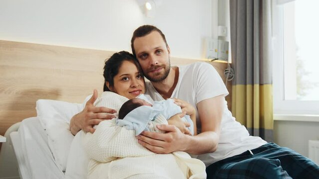 New mother and father in postnatal hospital department. Happy parents holding newborn baby boy in hospital ward. Smiling mother sitting in hospital bed with husband near and holding infant baby son