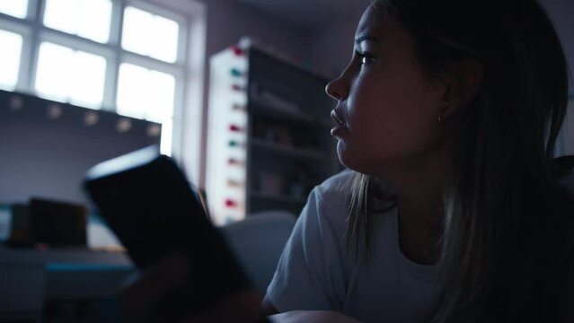 Depressed teenage girl lying on bed at home looking at bullying messages on mobile phone - shot in slow motion