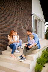Fototapeta premium Family with a mother, father, son and daughter sitting outside on the steps of a front porch of a brick house