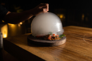 Wooden plate with Mexican food covered by bell with smoke on a wooden table with backlighting 