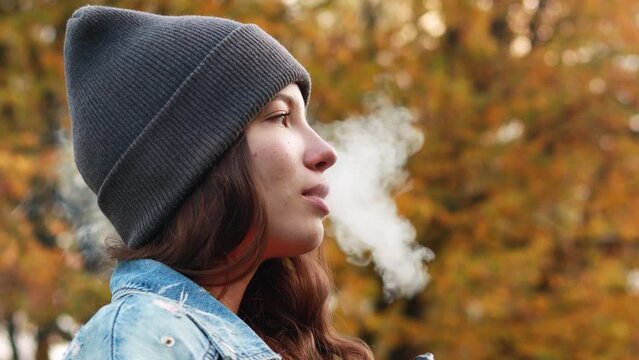 4k footage. Young woman in warm sunny autumn park season smoking tobacco device electronic cigarette heater vape. Smoke and steam system, fall, yellow orange red leaves
