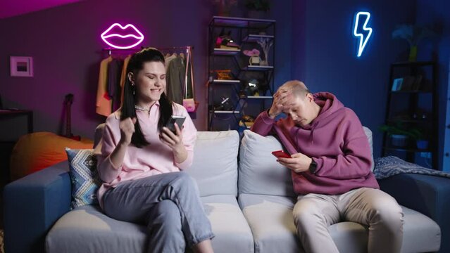 Romantic couple uses mobile phones. Man and woman play online game, gambling competitive game using phones. Woman rejoices in victory by dancing while loser man frustrated and aggressively angry.