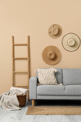 Cozy grey sofa with cushion, plaid in basket, ladder and hanging hats near beige wall