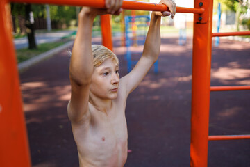 Fototapeta premium A teenager with an athletic development of musculature holds on to the horizontal bar. Street workout on a horizontal bar in the school park.