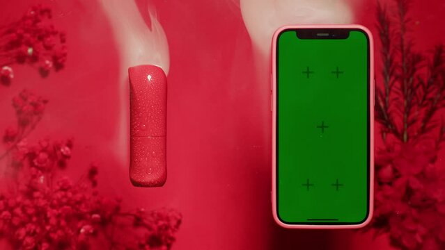 Phone with green screen chroma key on red and pink background close up, top view. Smartphone on summer bright texture studio shot. Composition of rose flowers, aroma and lipstick make