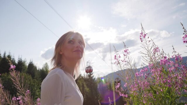 Young beautiful brunette woman walking through wildflowers in alpine meadow, summer dress, mountain, sunlight, wanderlust, happiness. 4K 24FPS