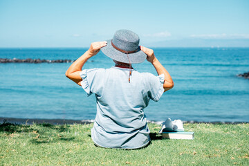 Back view of mature caucasian woman sitting in the meadow close the beach holding hat with hands looking at horizon over sea enjoying freedom and vacations