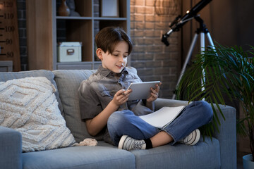 Little boy doing homework with tablet and sitting on sofa at home in evening