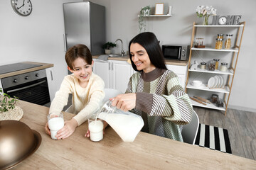 Happy little boy with his mother sitting at table and pouring fresh milk into glasses in kitchen