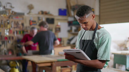 One young black apprentice of carpentry workshop taking notes on notepad, wearing apron and protective eye-wear. Staff employee engaged in learning business - obrazy, fototapety, plakaty